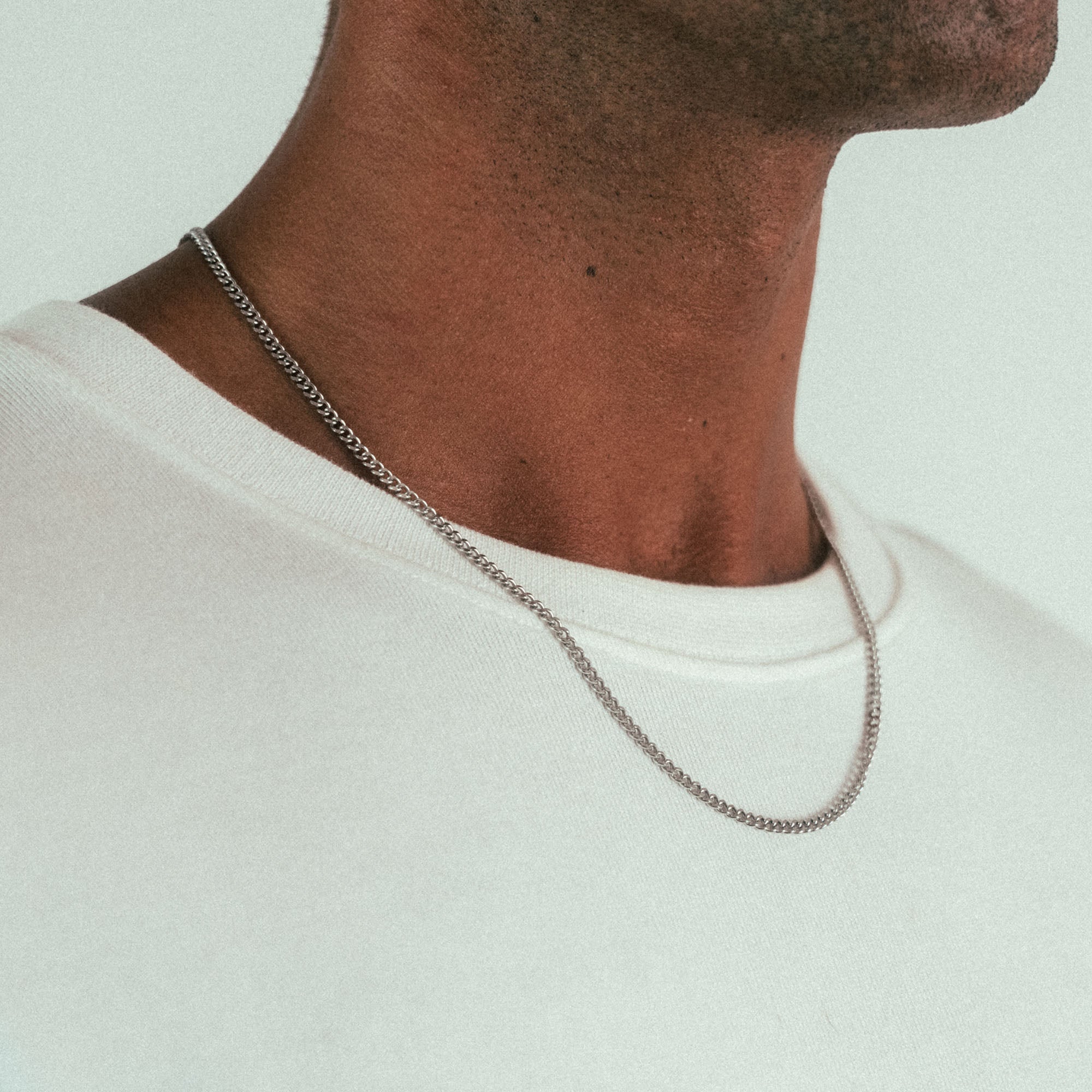 A close-up showcases someone wearing the WAV Cuban Chain Necklace elegantly over a light beige or white t-shirt. The silver chain gracefully drapes across the neck, highlighting the shoulder against a neutral background.