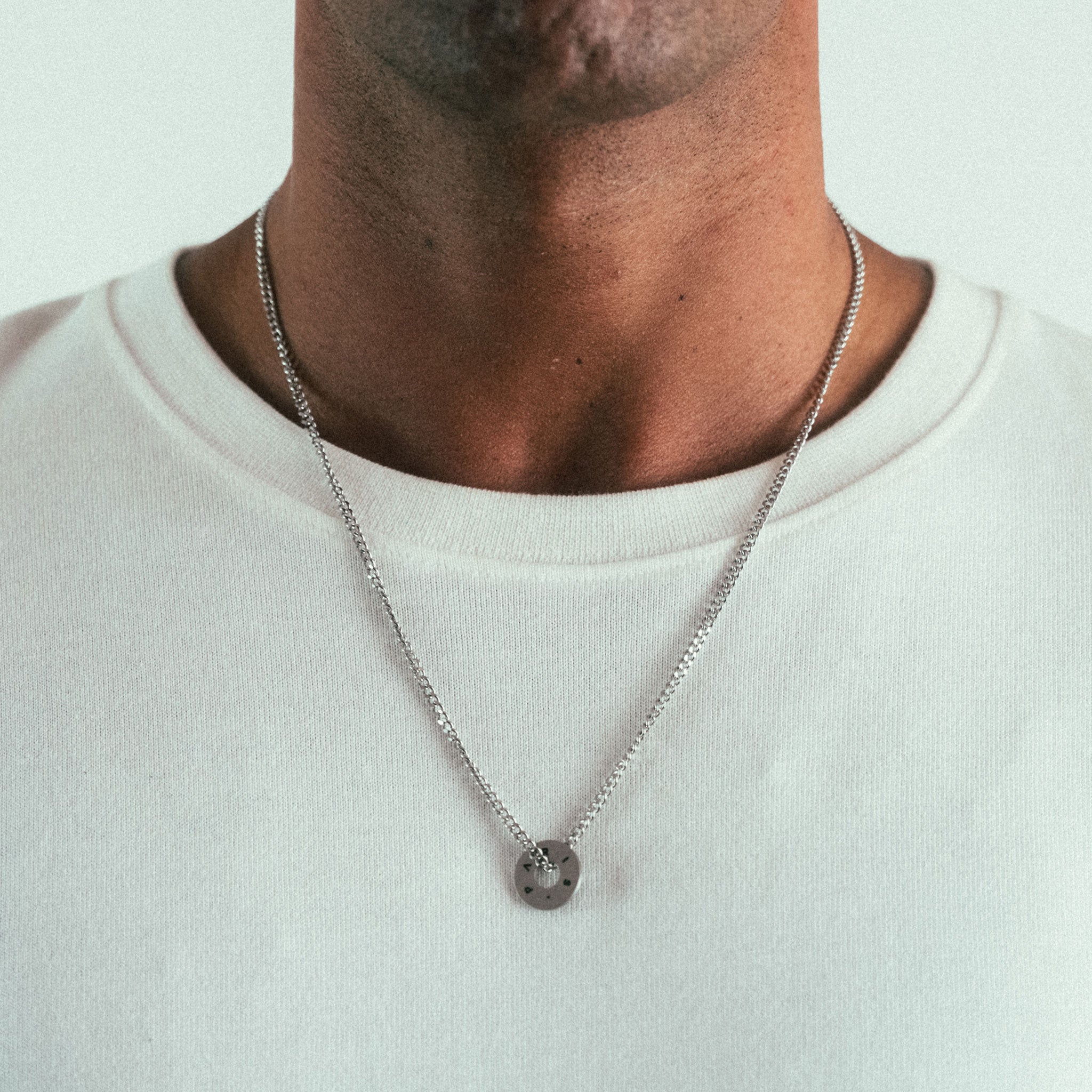A close-up shows a person wearing a plain white t-shirt and a WAV Custom Extra Ring silver chain necklace. The engraved pendant is simple and metallic against a soft, neutral background.