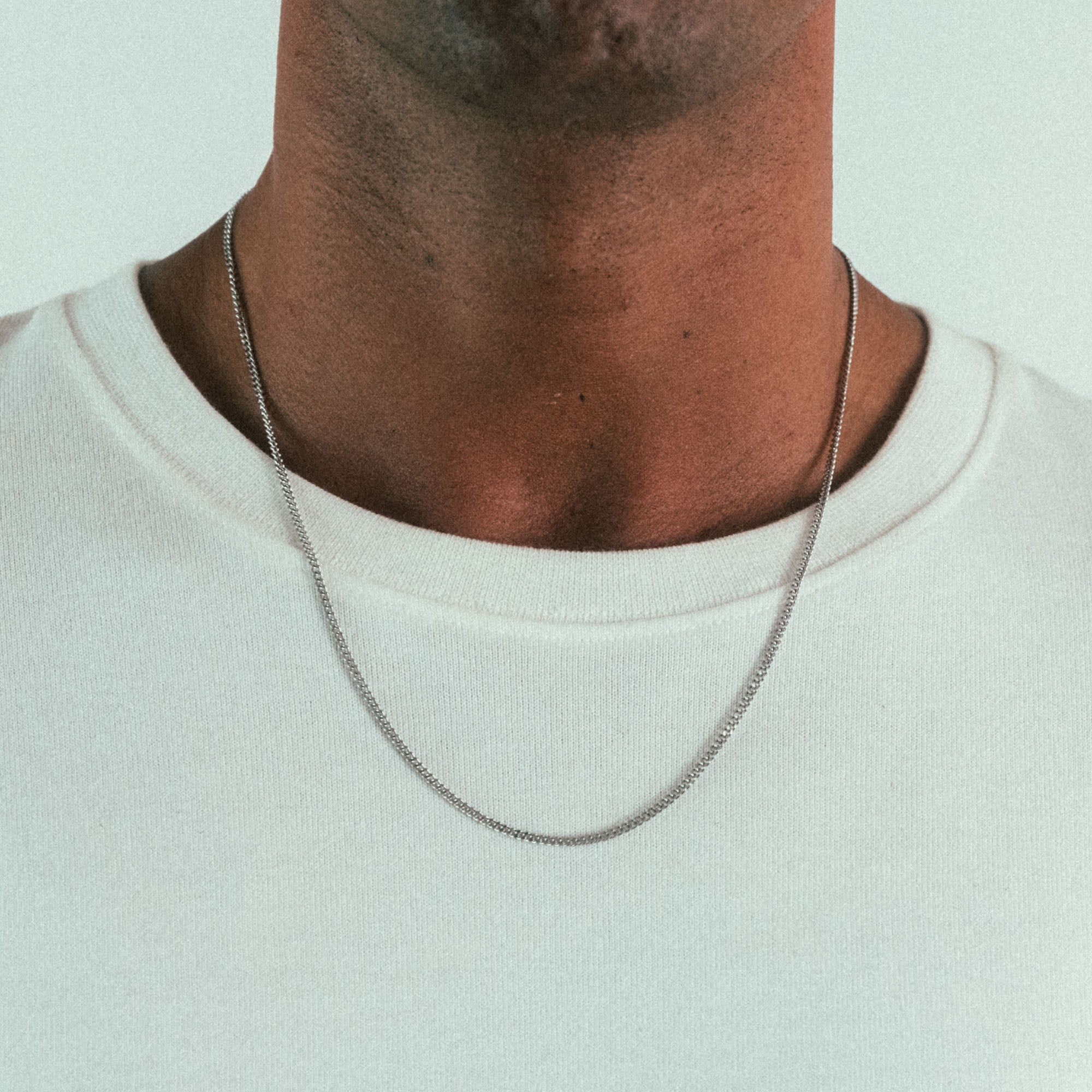 A close-up of a neck wearing WAVs Cuban Chain Necklace, a stylish piece of personalized jewelry over a plain white t-shirt, set against a solid light color background.