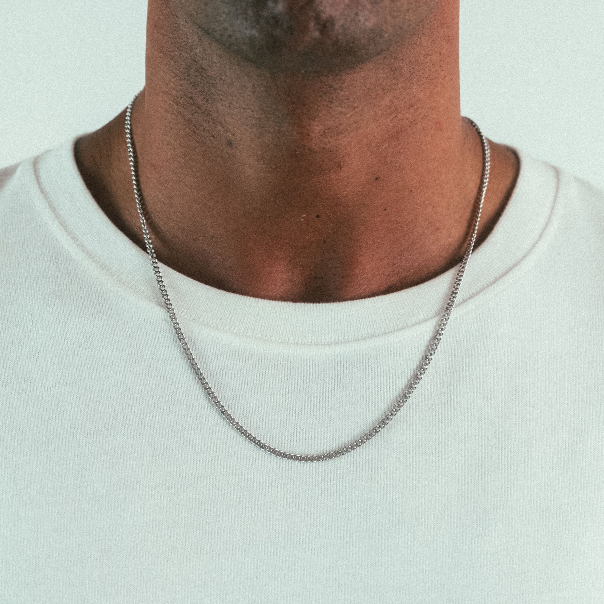 A person wearing a plain white t-shirt with a WAV Cuban Chain Necklace, highlighting personalized jewelry. The shot focuses on the neck and upper chest against a plain, neutral background.