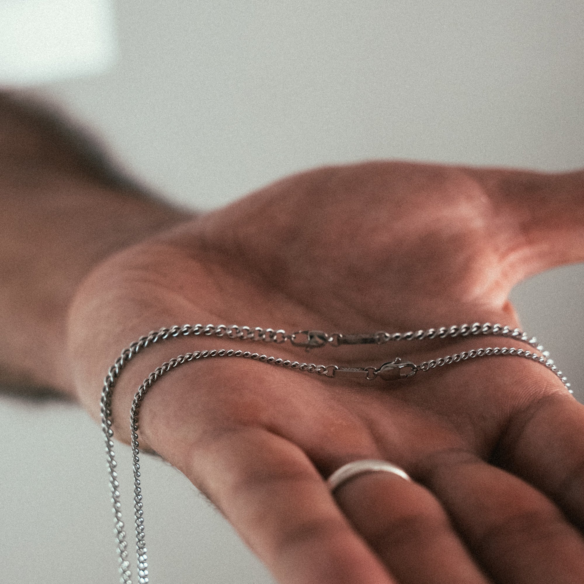 A persons hand holds a delicate, broken silver chain with a clasp, showcasing the craftsmanship of WAVs Custom Necklace Ring. The hand is slightly angled, adorned with a silver ring on the pinky finger. A blurred neutral background enhances the scene, imbuing it with the feeling of receiving cherished jewelry.