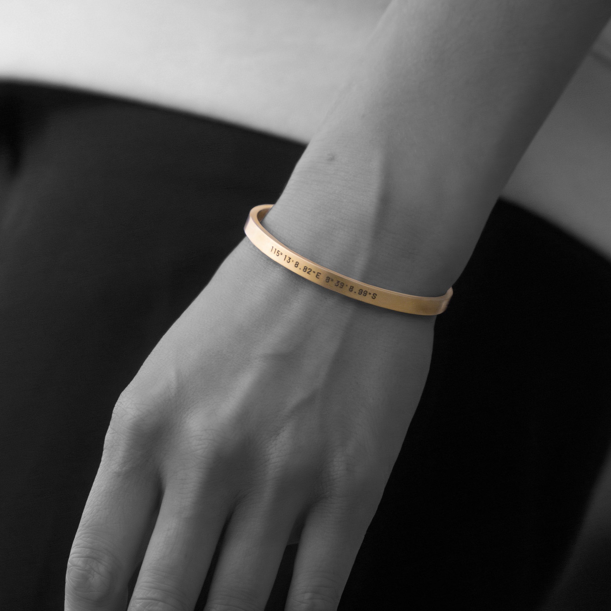 Close-up of a persons left hand wearing the gold Custom Cuff Bracelet by WAV, highlighting personalized jewelry. The image is mainly black and white, with the bracelet in gold to emphasize its presence.