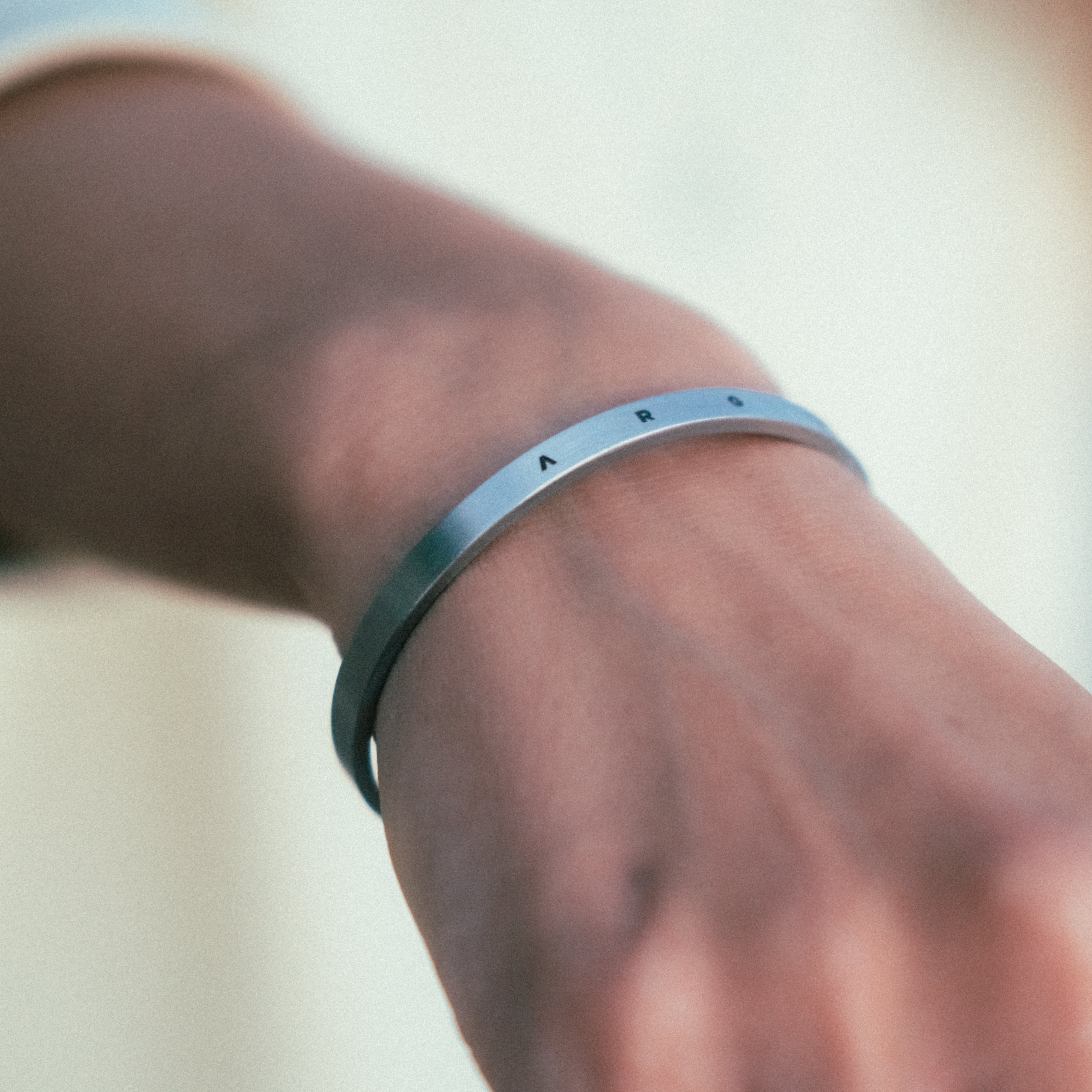 A close-up shows a wrist wearing WAVs sleek metallic Custom Cuff + Necklace Bundle, engraved with A R. The blurred background accentuates the minimalist elegance of this custom jewelry piece.