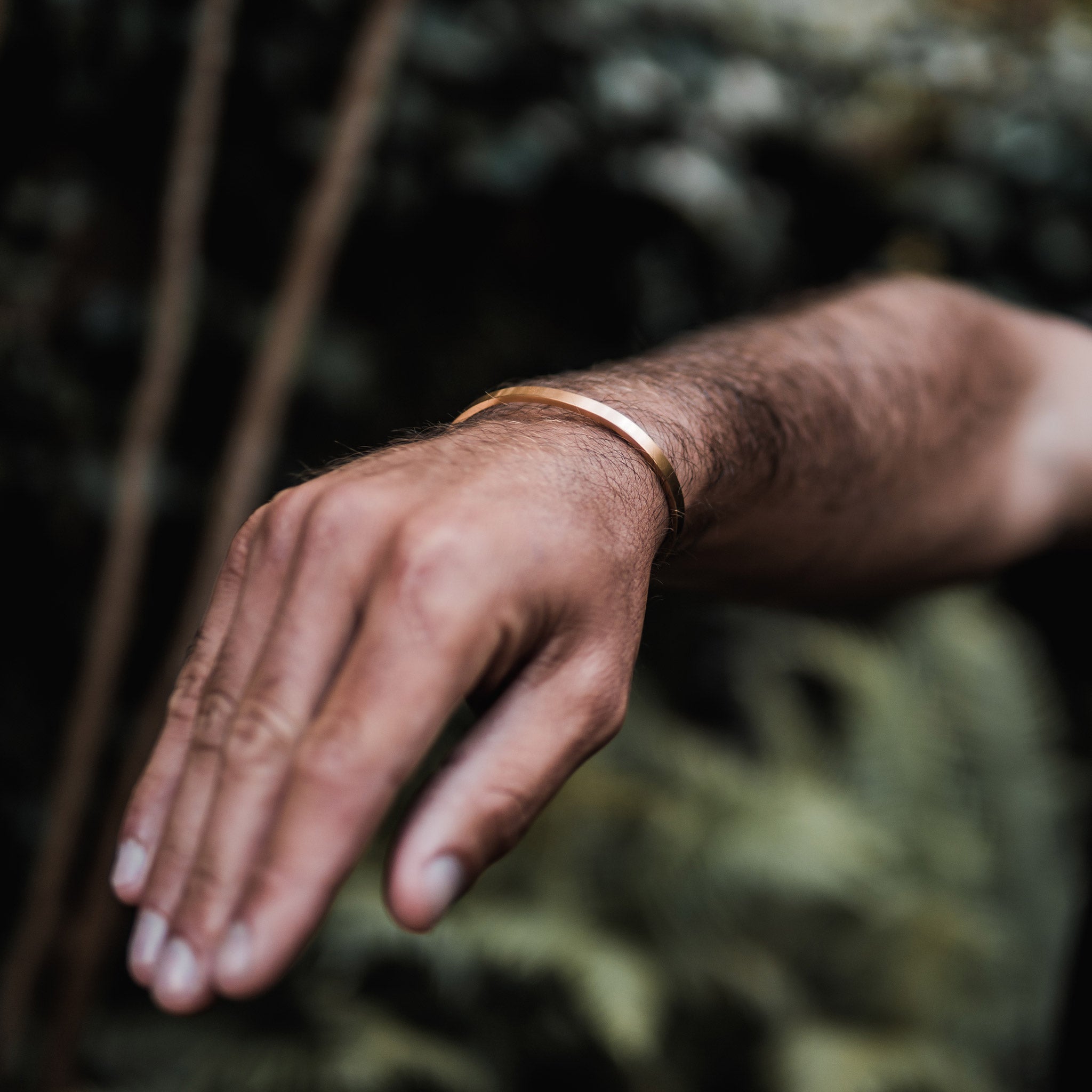Custom cuff bracelet on a man's wrist, made of stainless steel 316L with 18k gold PVD plating, customized and personalized with Morse code, coordinates, names, initials, dates, or custom text. Minimalist unisex jewelry, displayed against a natural, green background
