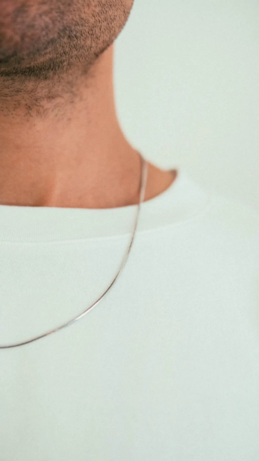 A close-up of a persons neck with light stubble, wearing the WAV Mamba Necklace—a thin silver chain—paired with a white crew-neck shirt against a plain light background.