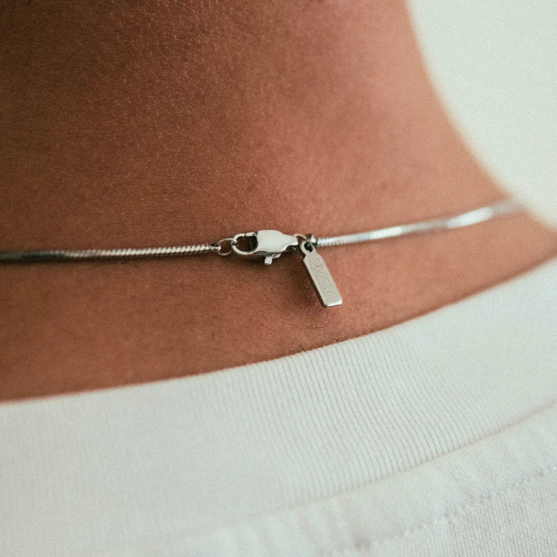A close-up of the back of a person’s neck wearing the WAV Mamba Necklace—a thin, minimalist piece with a clasp and small rectangular tag—paired with a white shirt.