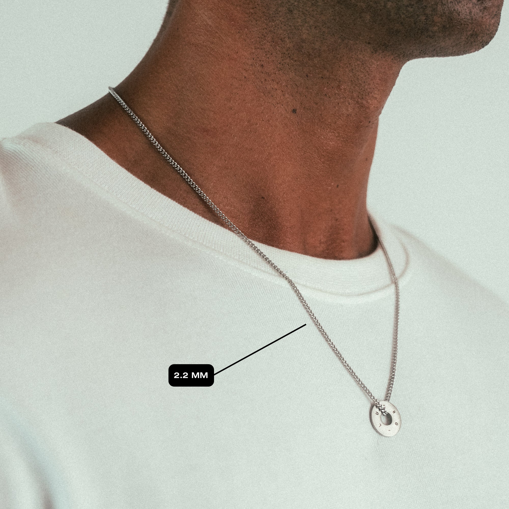 Close-up of a person wearing the Custom Necklace Ring by WAV, a silver chain with a round pendant over a white t-shirt. A label points to it with 2.2 MM, indicating its thickness, showcasing exquisite custom jewelry.