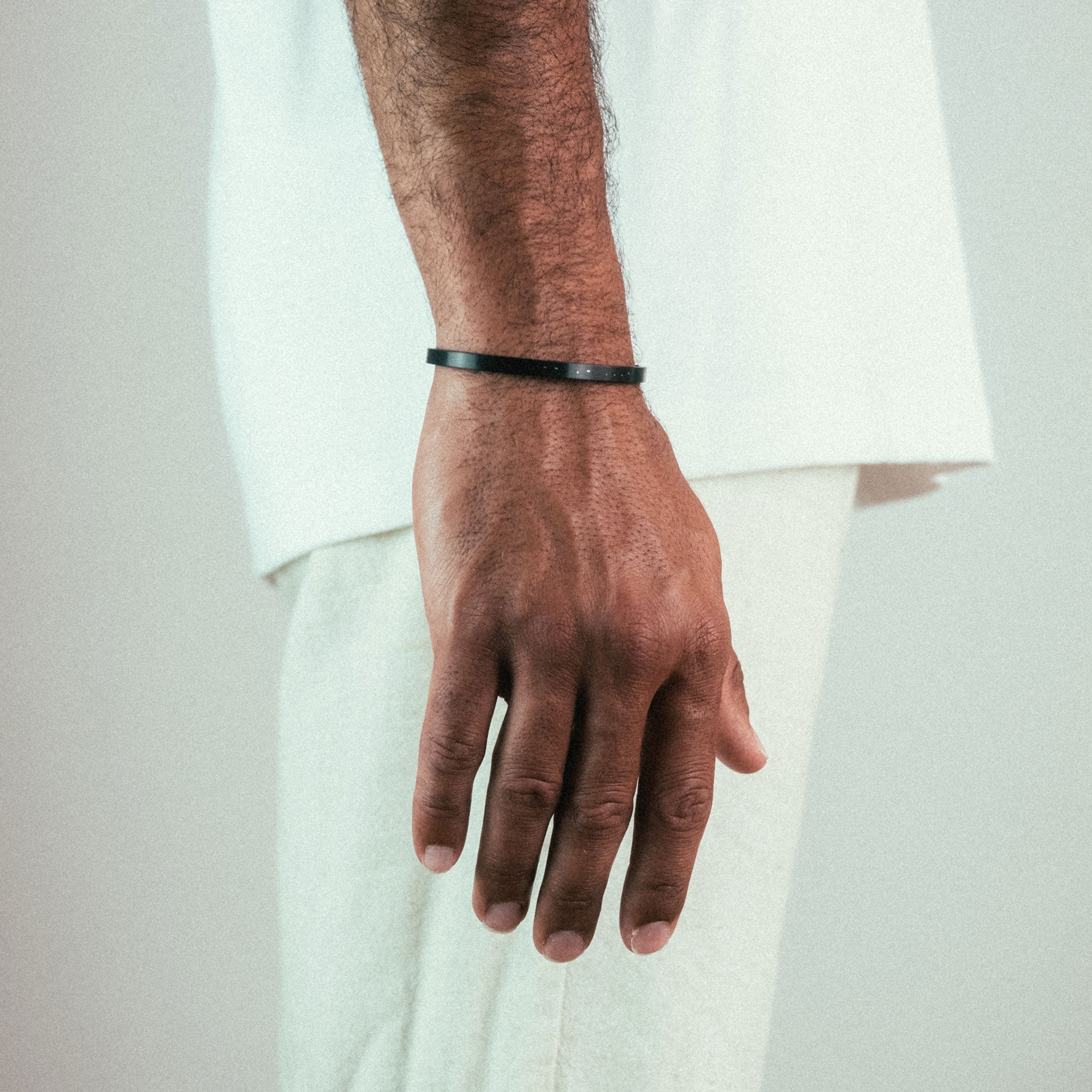A close-up shows a person wearing WAVs thin black Custom Cuff Bracelet, subtly personalized. They wear a light short-sleeve shirt and pants against a neutral background.