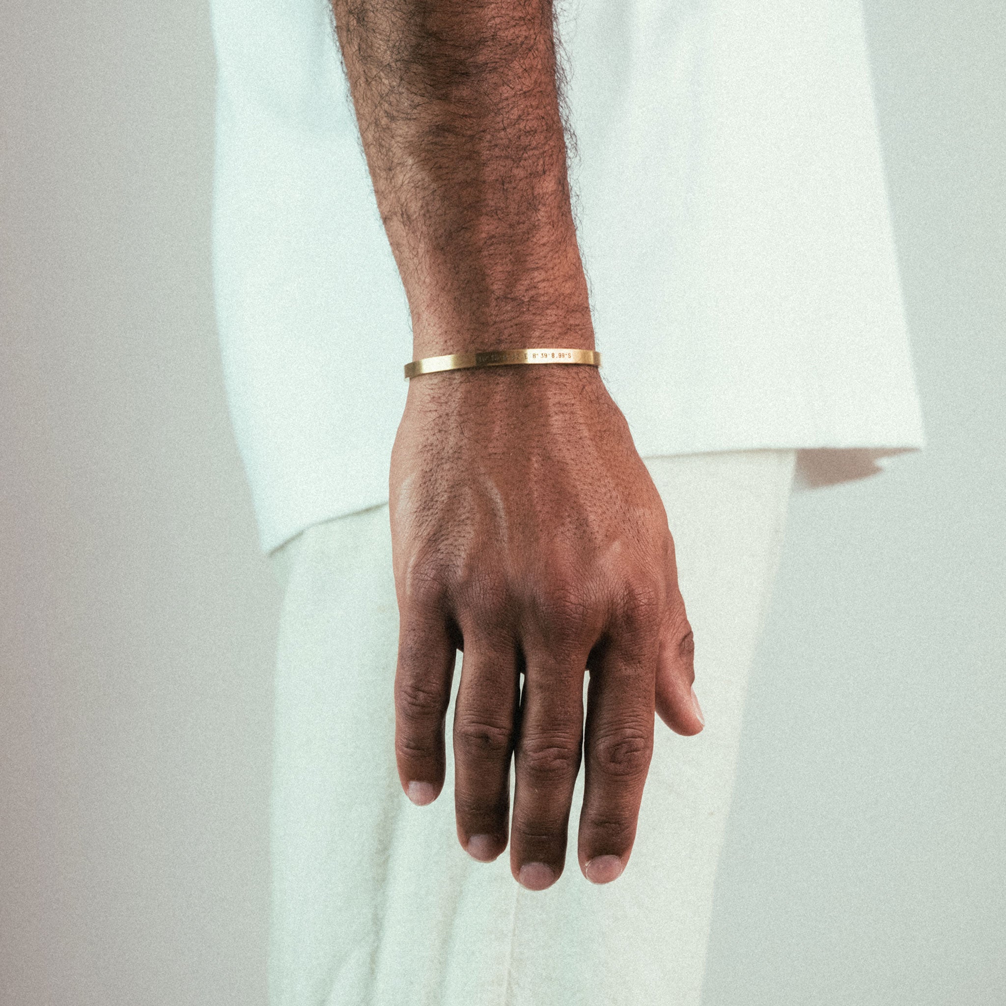 A close-up of a hand wearing an engraved WAV Custom Cuff Bracelet, the gold tone shining against the medium-dark skin and white outfit. The neutral background enhances the focus on this personalized jewelry piece.
