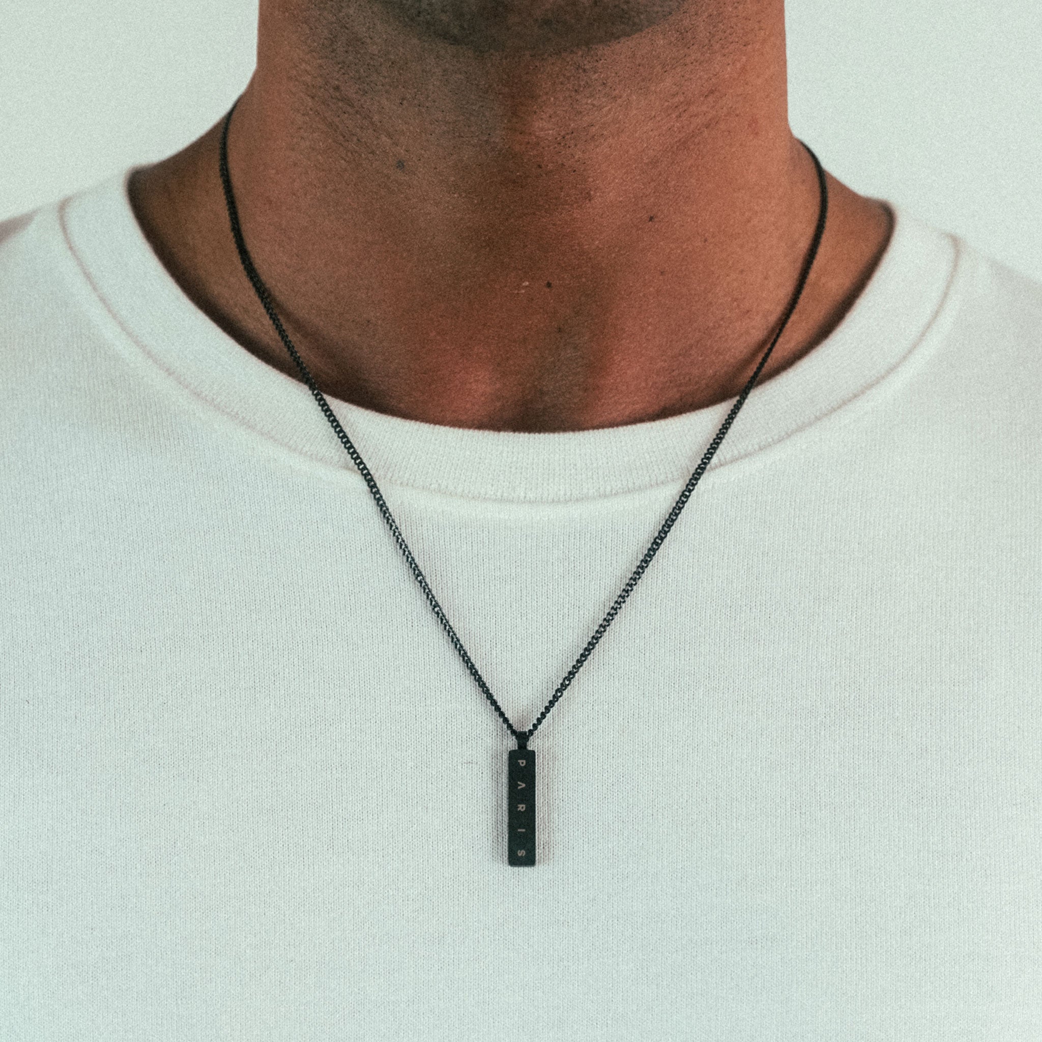 A person with a plain white T-shirt wears a WAV Custom Necklace Bar, which features embossed markings for added sophistication. The focus is on the neckline and personalized jewelry, highlighting its unique charm.