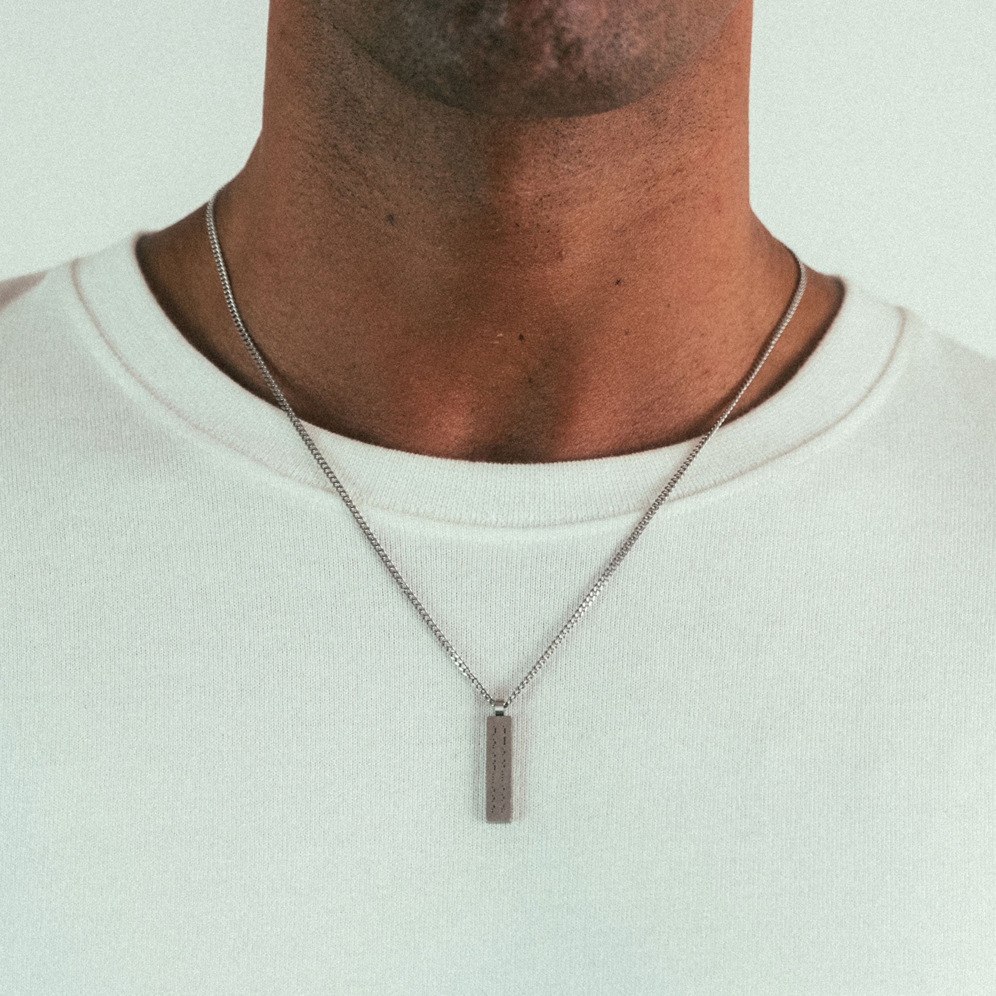 A person in a white shirt, shown from shoulders to above the mouth, sports a silver WAV Custom Necklace Bar with an elegantly engraved pendant. The neutral, out-of-focus background highlights this exquisite personalized jewelry piece.