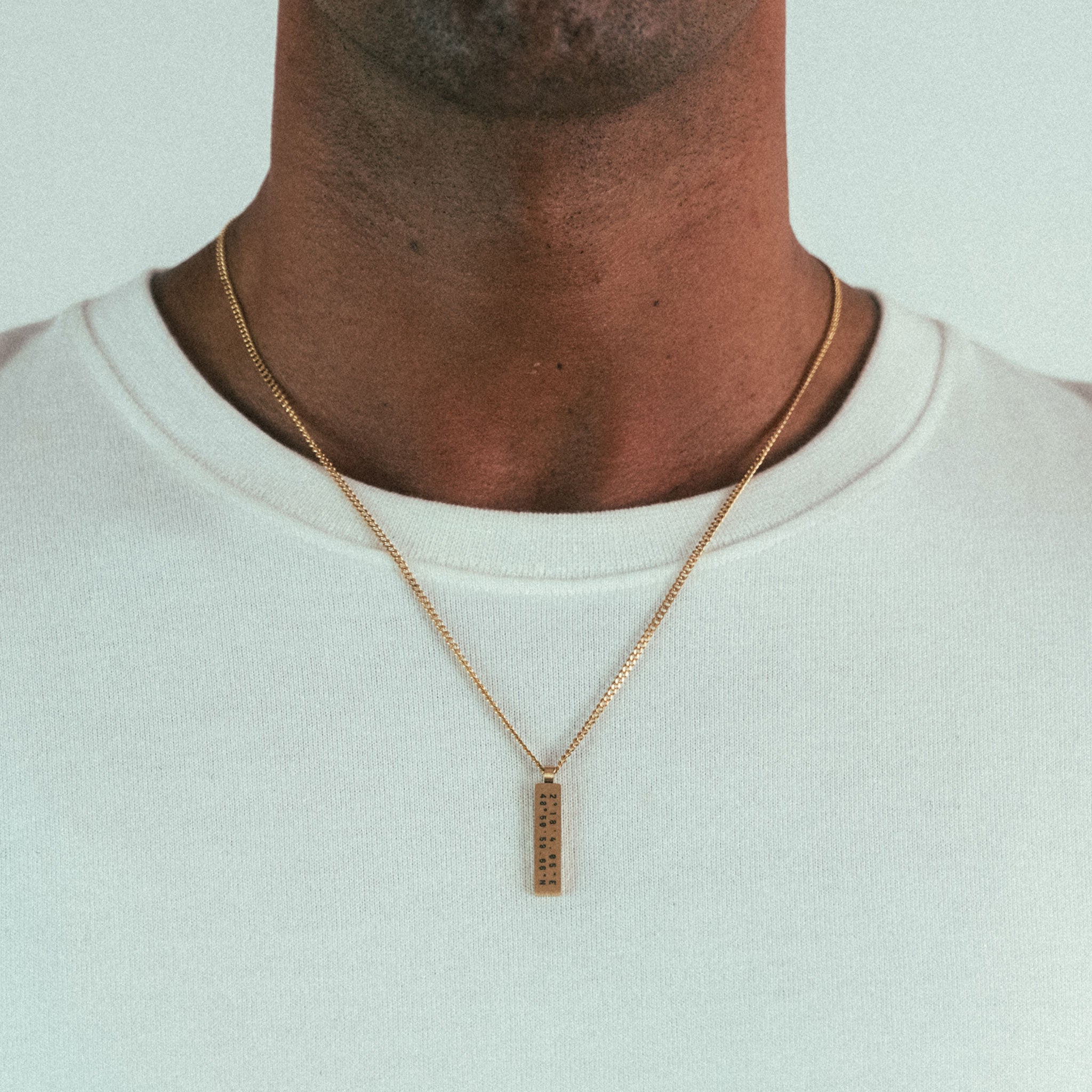 A person wears a WAV Custom Necklace Bar with a gold chain, featuring engraved patterns on its rectangular pendant. The photo highlights the personalized custom jewelry on their white shirt and upper chest area.
