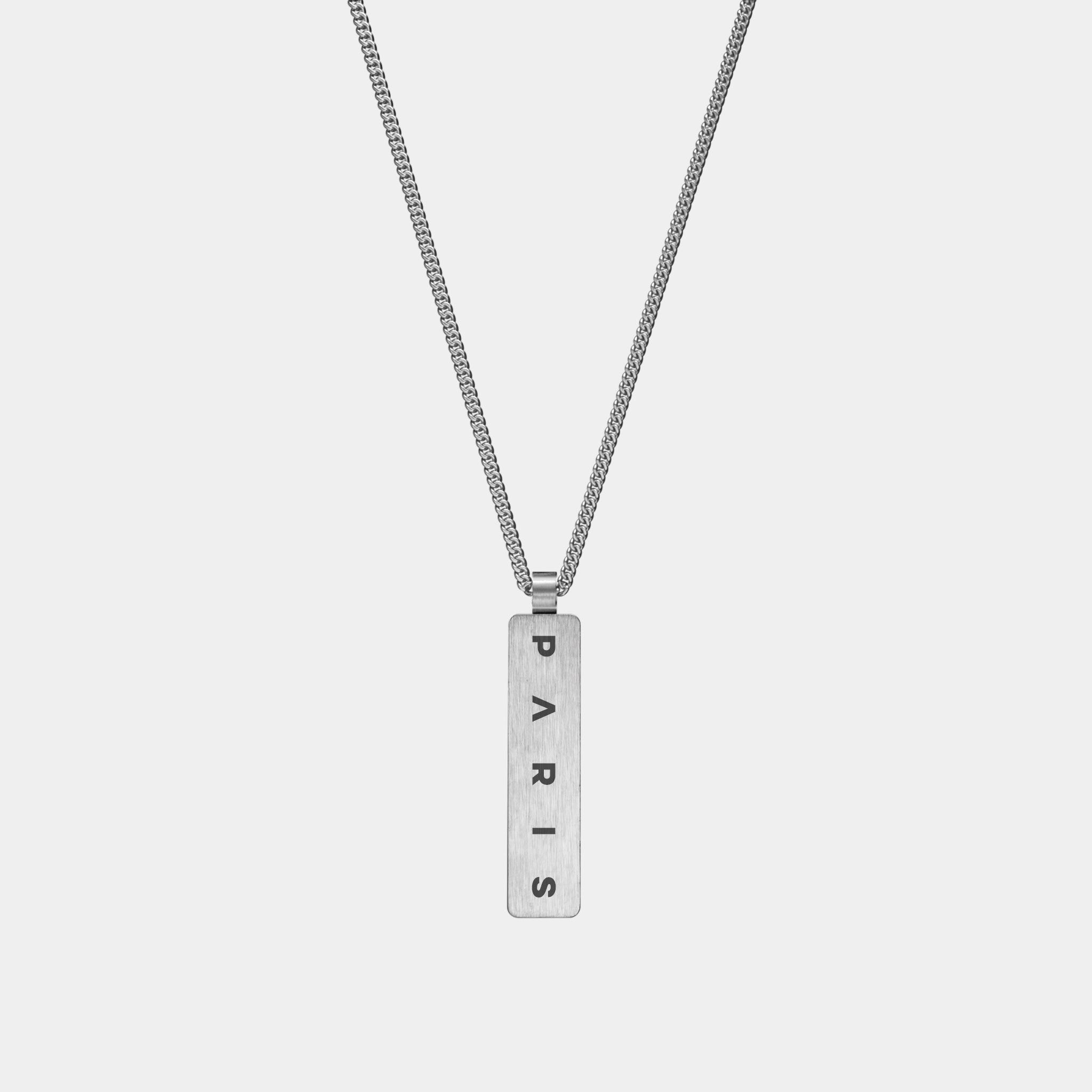 The WAV Custom Necklace Bar is a silver pendant with a vertical rectangular tag engraved with PARIS in black capitals on a simple, thin link chain, embodying the elegance of personalized jewelry against a plain white background.