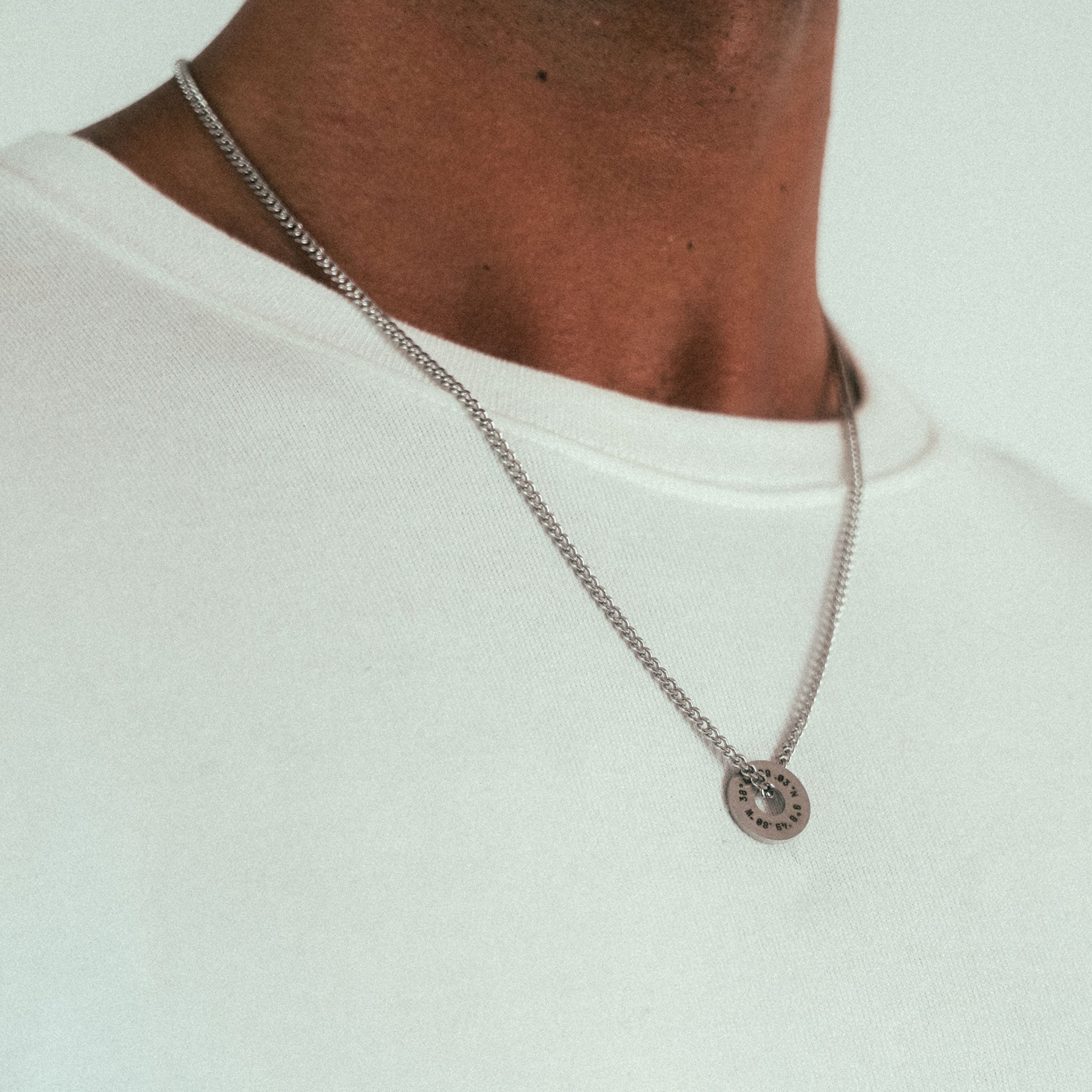 A person wears a plain white shirt paired with the WAV Custom Extra Ring necklace featuring a silver round pendant with intricate engraving. The shot highlights their upper torso and neck against a neutral, light background.