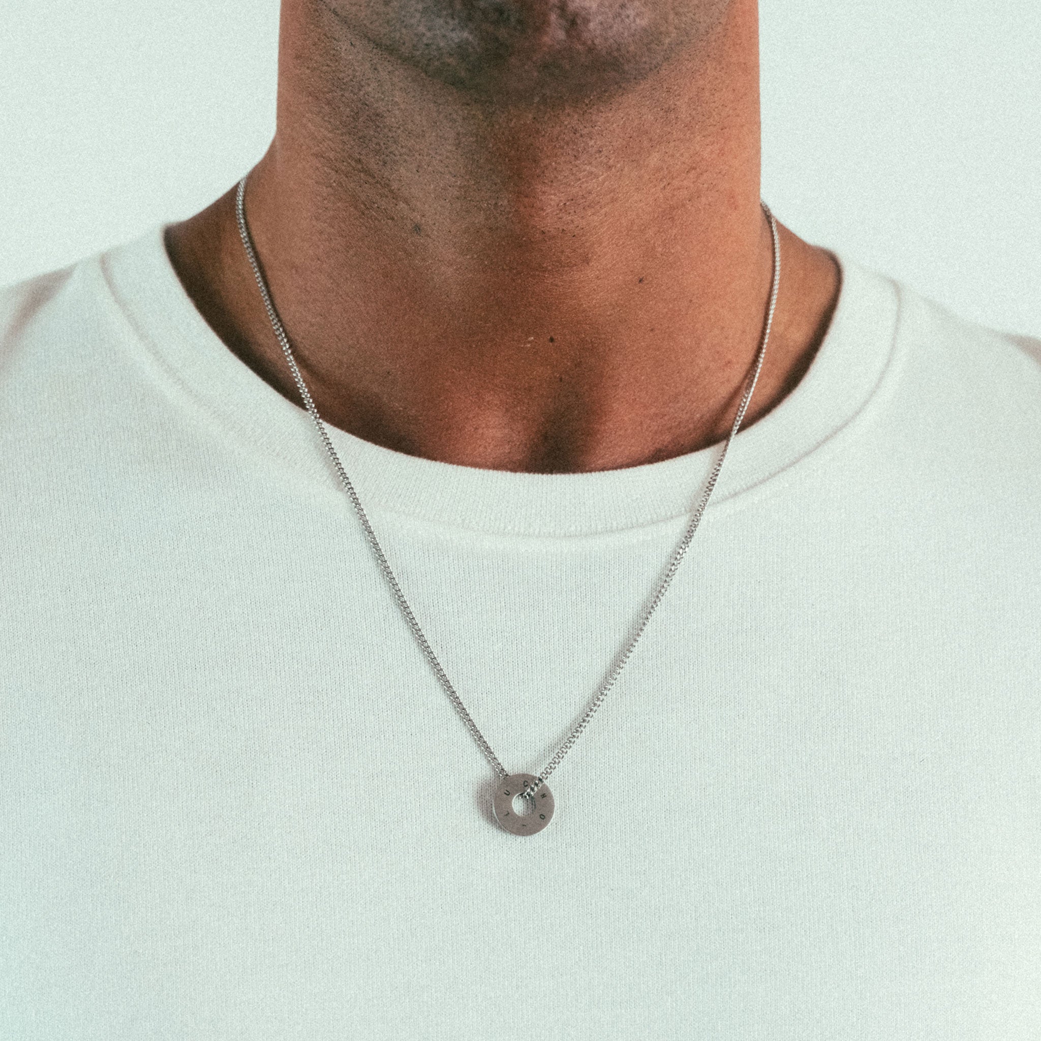 A person dons a WAV Custom Necklace Ring with a simple silver chain and round pendant, elegantly resting on their white t-shirt, highlighting the personalized jewelry against the plain shirt.