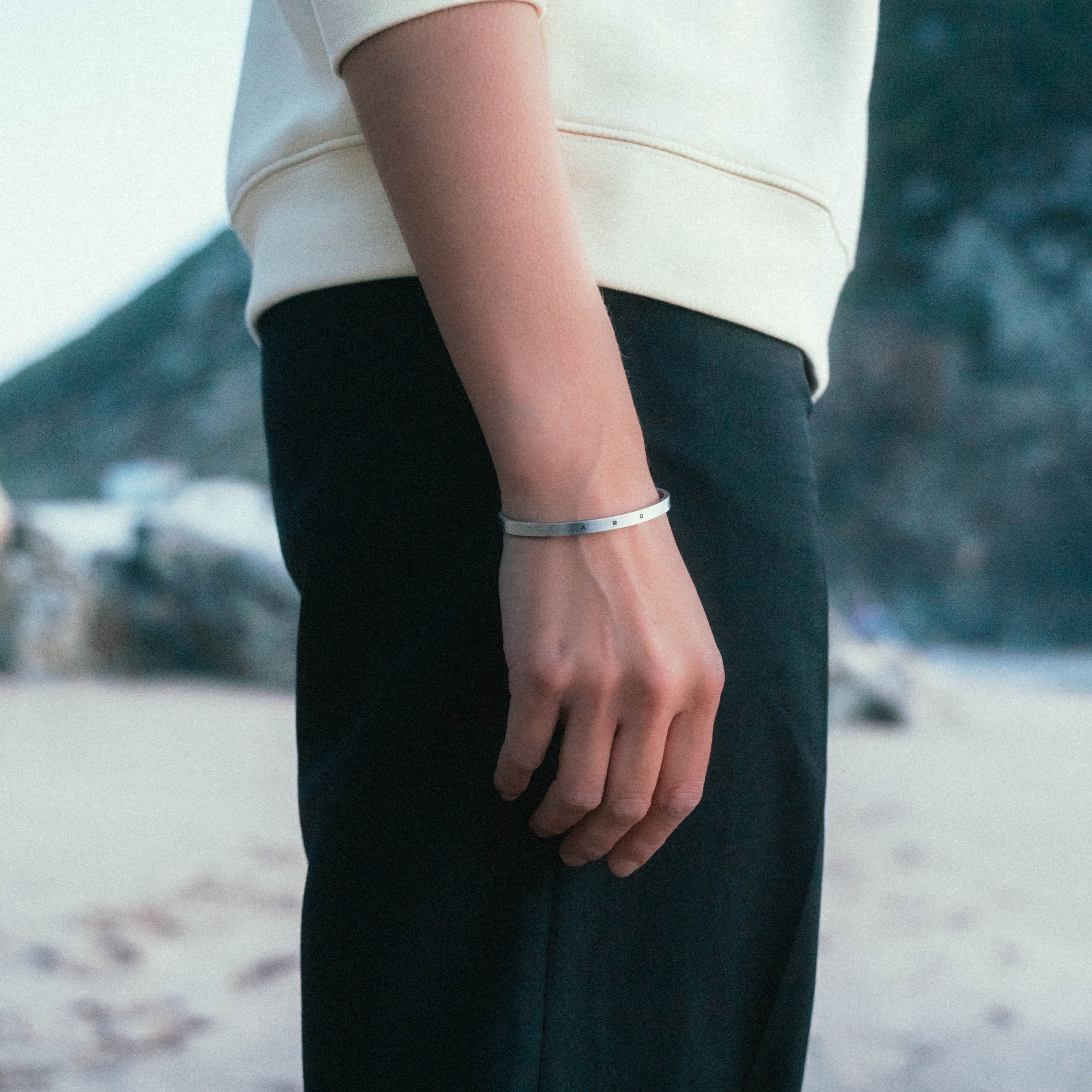 A person stands on a beach with mountains in the background, wearing dark pants, a light long-sleeve shirt, and a Custom Cuff Bracelet by WAV. The serene scene highlights their hand and bracelet, adding an aura of calmness.